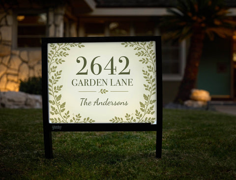 Illuminated address yard sign showing house number, street and family name at dusk
